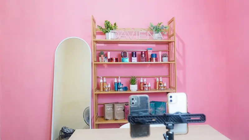 Tampilan horizontal studio berlatar dinding merah muda, rak produk dan tanaman hias, cermin tinggi, serta dua ponsel pada dudukan di depan meja.