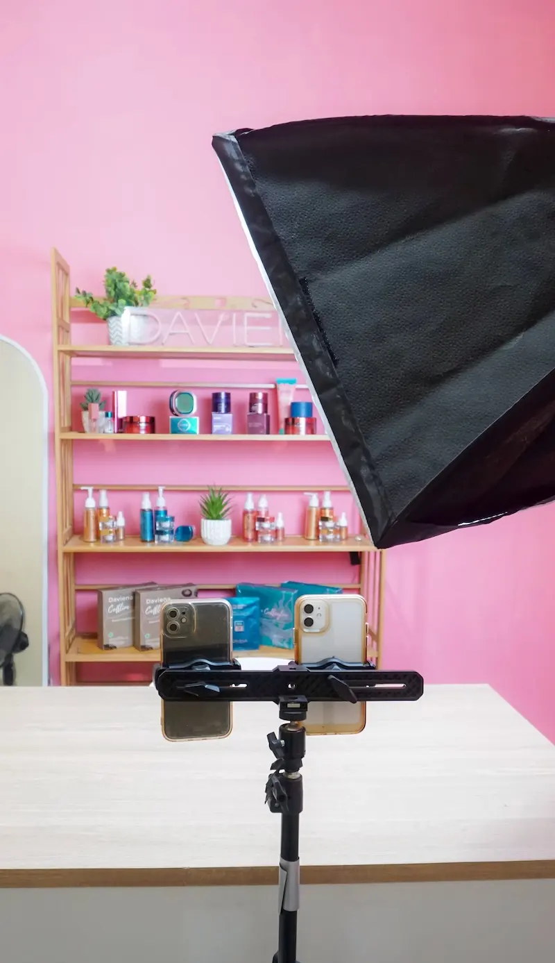 Tampilan vertikal studio produk berwarna merah muda dengan dua ponsel pada tripod, meja putih, rak produk, dan softbox hitam di kanan atas.