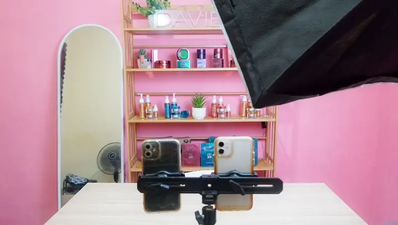 Dua ponsel pada dudukan horizontal di depan meja putih, dengan rak produk, cermin, dinding merah muda, dan sebagian softbox di latar.