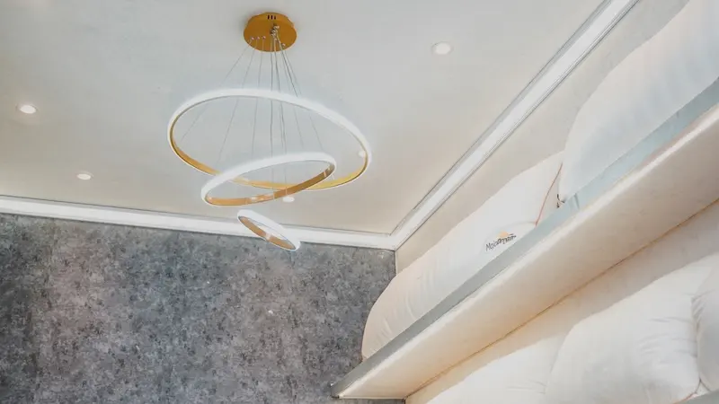 Detail langit-langit ruangan dengan lampu gantung berbentuk cincin, dinding bertekstur abu-abu, serta rak bantal putih di sisi kanan.