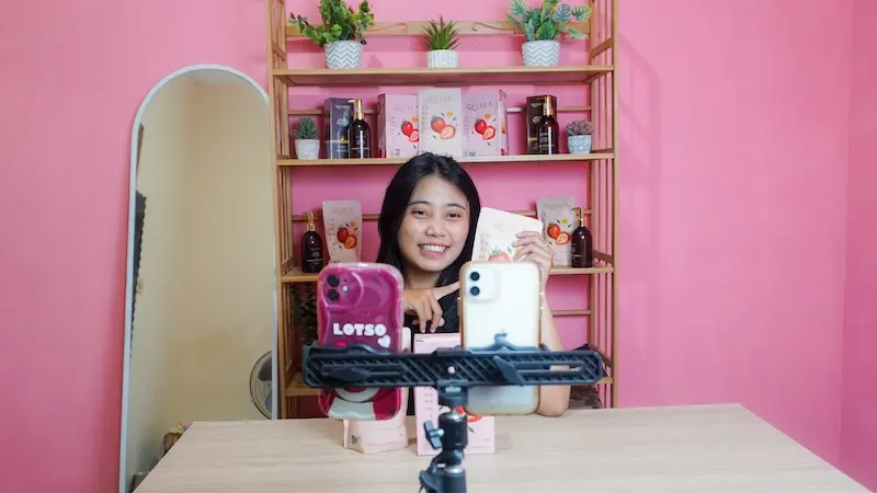 Host memperlihatkan produk kemasan merah muda di meja studio, dengan dua ponsel di depannya.