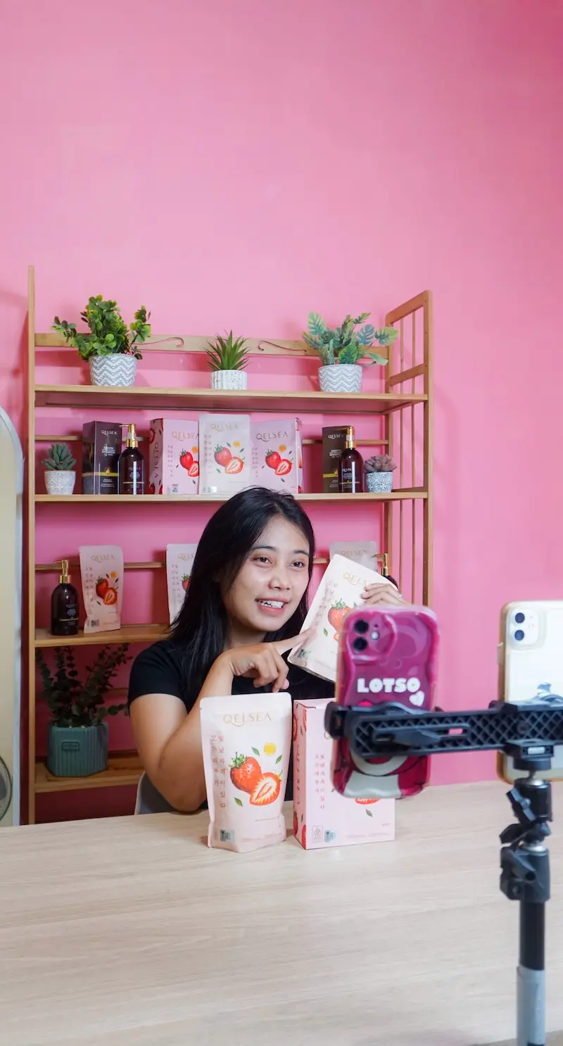 Tampilan vertikal seorang perempuan di meja studio merah muda, dengan kotak produk di meja serta ponsel pada dudukan di depan.