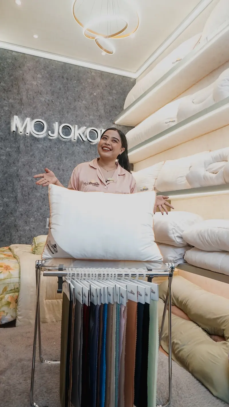 Seorang perempuan berdiri sambil memegang bantal putih di ruang pajangan perlengkapan tidur; rak bantal dan sampel kain terlihat di sekelilingnya.