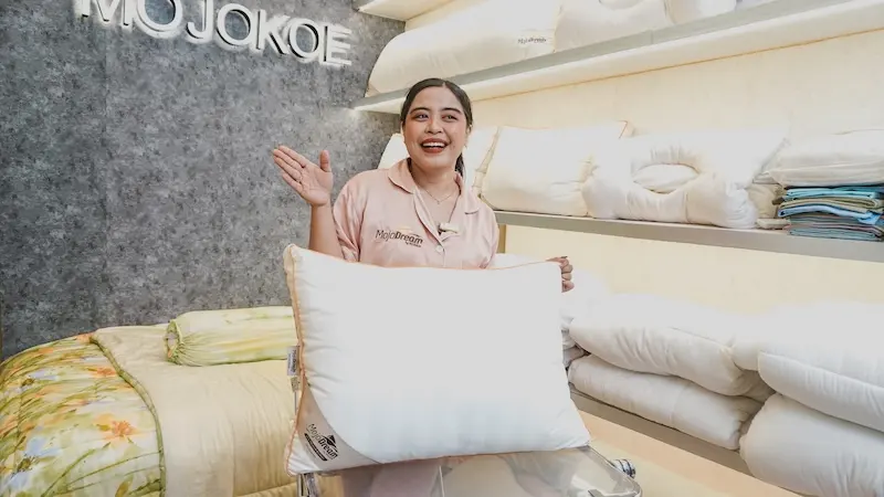 Tampilan horizontal seorang perempuan memperagakan bantal putih dan mengangkat satu tangan, dengan rak bantal dan dinding MOJOKOE di belakang.