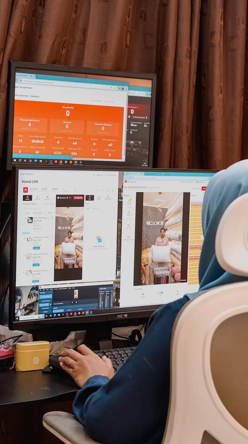 Seseorang berhijab biru duduk di kursi putih mengoperasikan komputer dengan dua monitor yang menampilkan halaman dan dasbor.