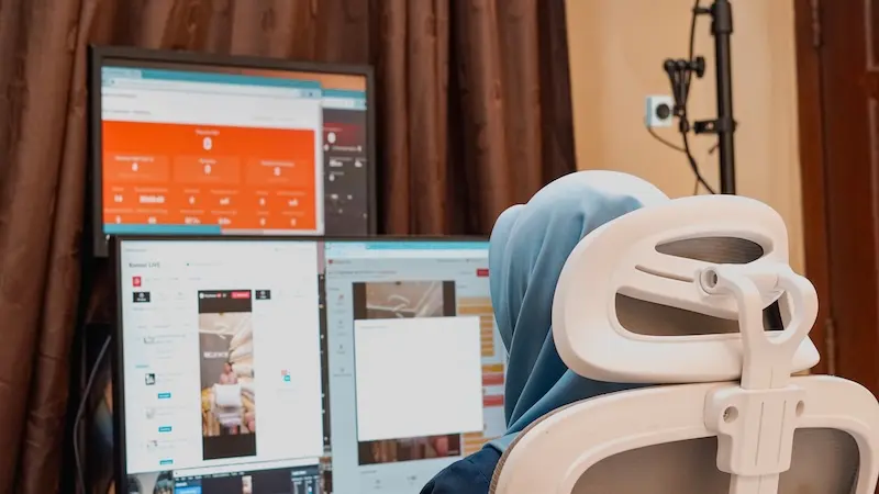 Area kerja operator berhijab biru dengan dua monitor, kursi kantor putih, meja, dan tirai cokelat di belakang.