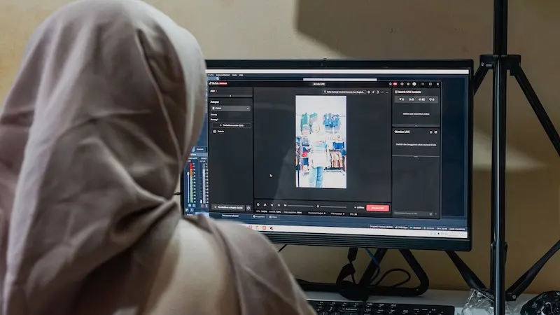 Seseorang berhijab abu-abu terlihat dari belakang menghadap monitor yang menampilkan antarmuka produksi video dengan pratinjau vertikal.