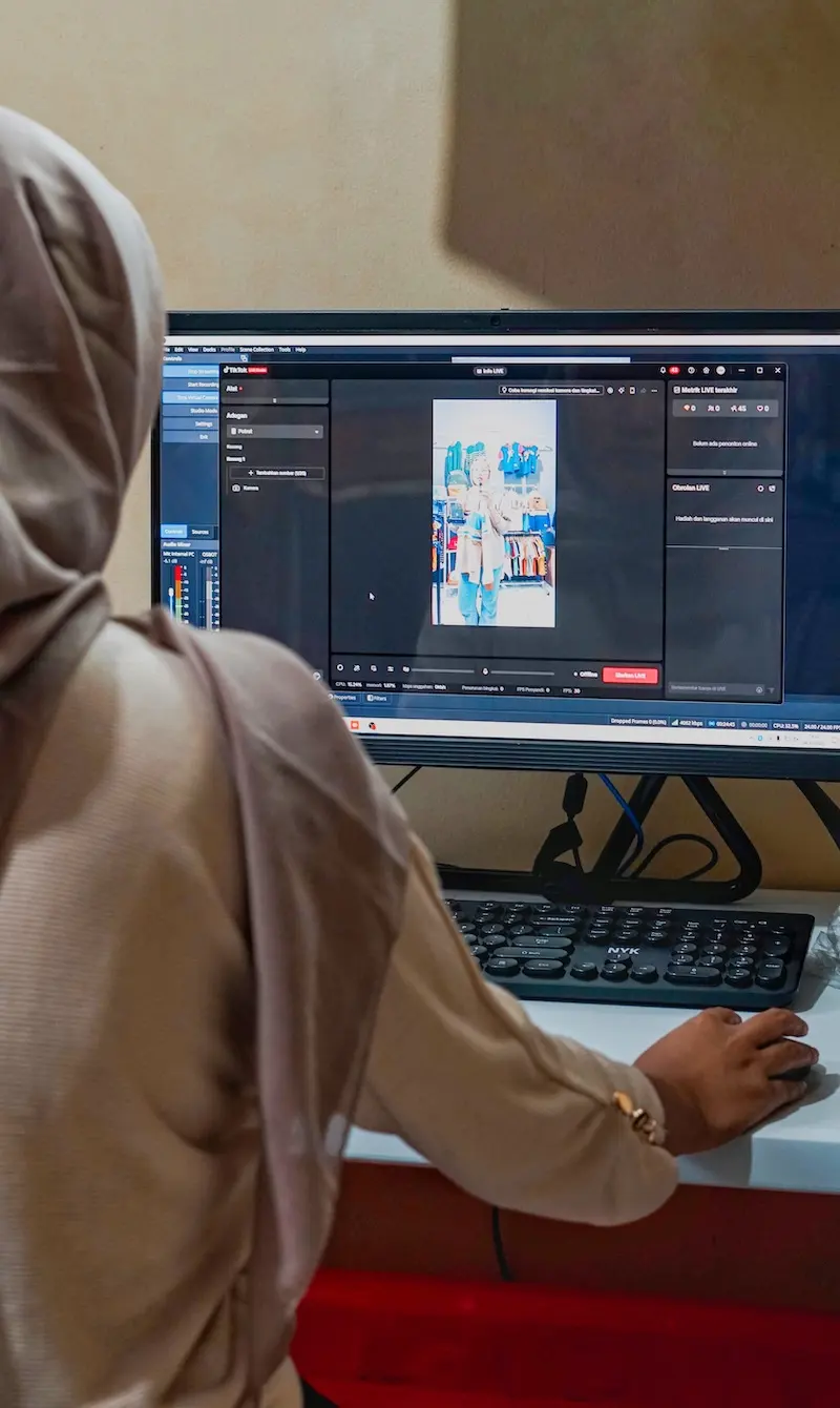 Tampilan vertikal operator berhijab abu-abu menggunakan komputer dengan pratinjau video vertikal pada layar.