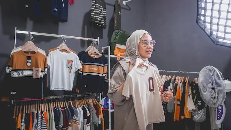 Seorang perempuan berhijab dan berkacamata berdiri di studio pakaian, dengan gantungan baju, kipas angin, dan lampu softbox.