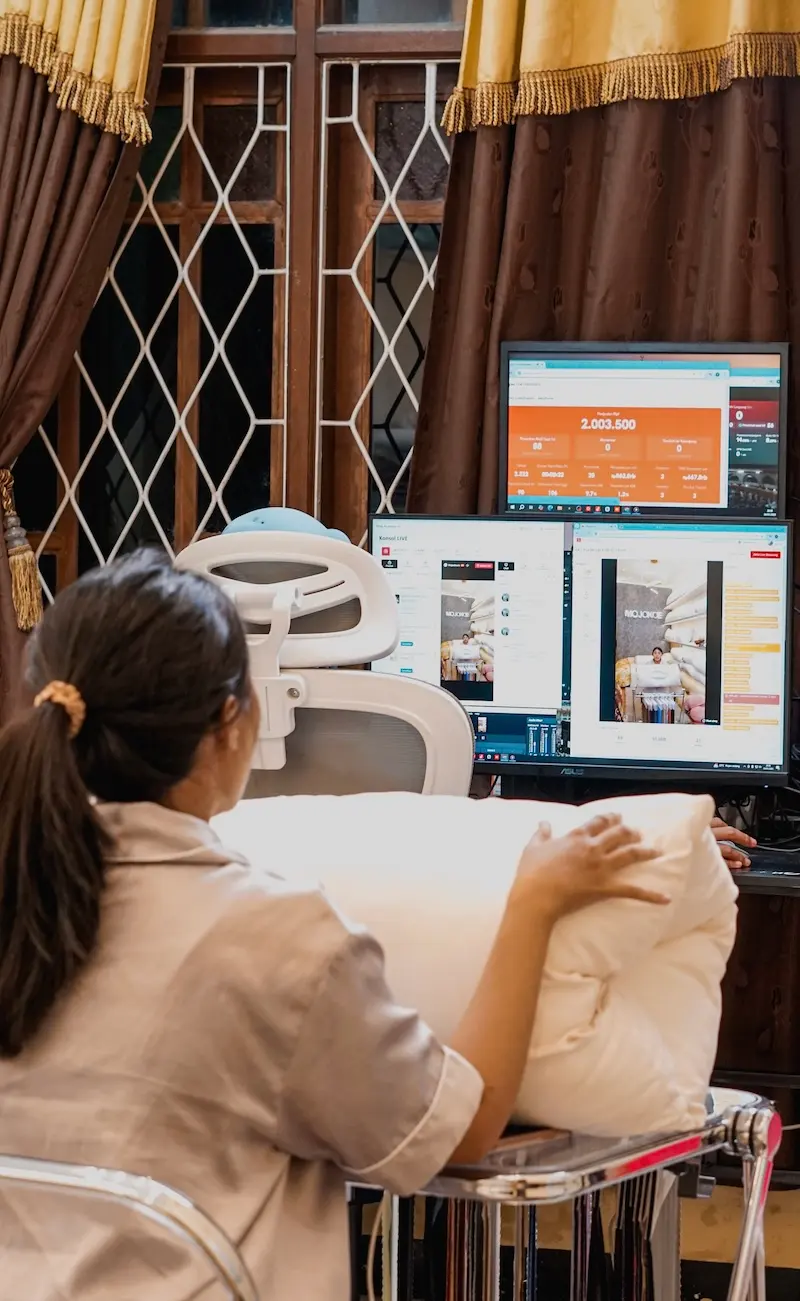 Seorang perempuan memegang bantal putih di dekat meja operator dengan dua monitor, kursi putih, dan tirai cokelat.