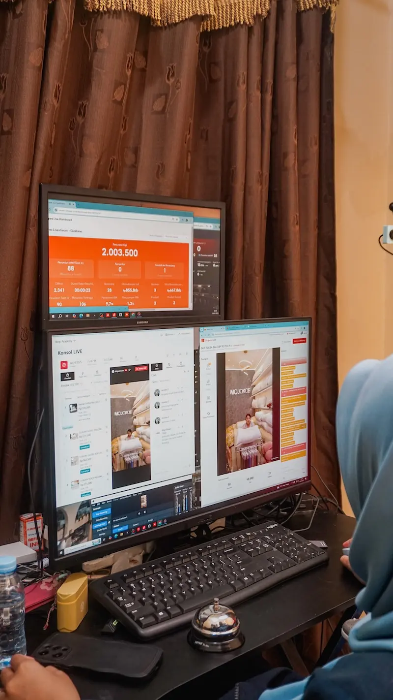 Tampilan vertikal meja operator dengan dua monitor bertumpuk yang menampilkan dasbor dan halaman pratinjau, keyboard, serta tirai cokelat.