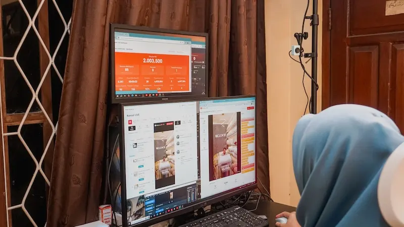 Tampilan horizontal operator berhijab biru di depan dua monitor bertumpuk, dengan tirai cokelat dan jendela di belakang.