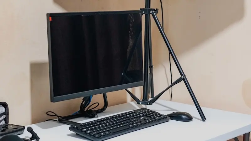 Monitor dengan layar gelap, keyboard, dan mouse di meja putih; kaki penyangga lampu berdiri di sampingnya.