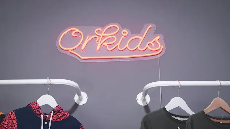Tulisan neon Orkids berwarna merah muda pada dinding gelap di atas gantungan pakaian.