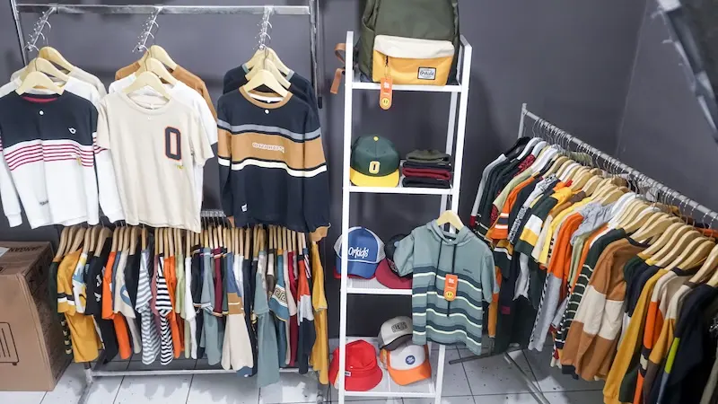 Sudut lebih dekat rak pakaian berisi beragam atasan pada hanger kayu, dengan tas dan topi pada rak tengah.