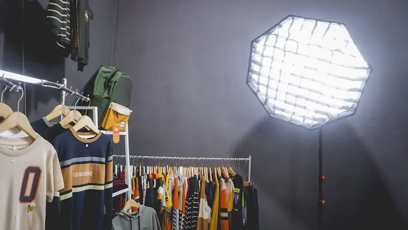 Lampu softbox segi delapan dengan kisi yang menyala di studio pakaian berdinding gelap, dengan rak pakaian di bawahnya.
