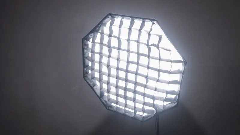 Foto dekat lampu softbox segi delapan dengan kisi hitam yang menyala terang pada tiang penyangga.