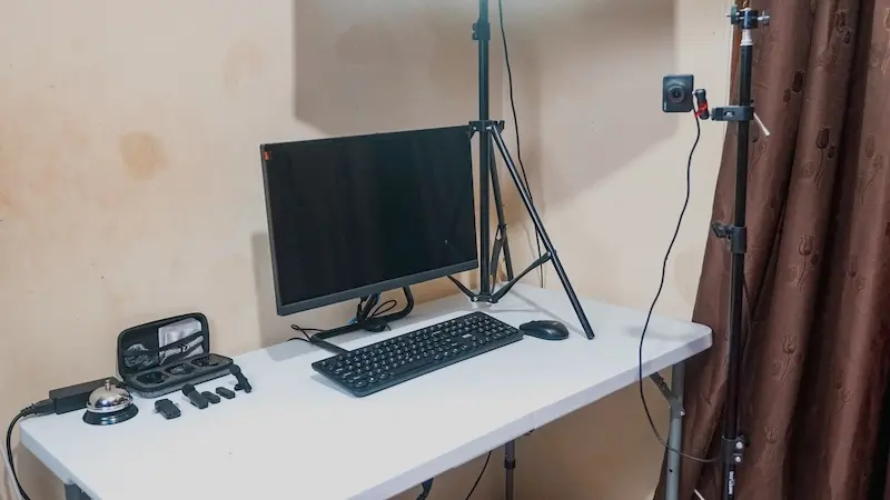 Tampilan meja peralatan studio dengan monitor gelap, keyboard, perangkat kecil, tripod ponsel, dan penyangga lampu di dekat tirai.