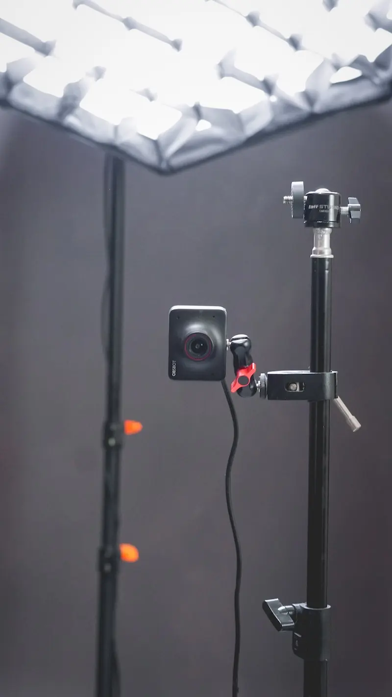 Tampilan vertikal kamera kecil pada dudukan dan tiang penyangga di bawah lampu studio yang menyala, dengan dinding gelap sebagai latar.