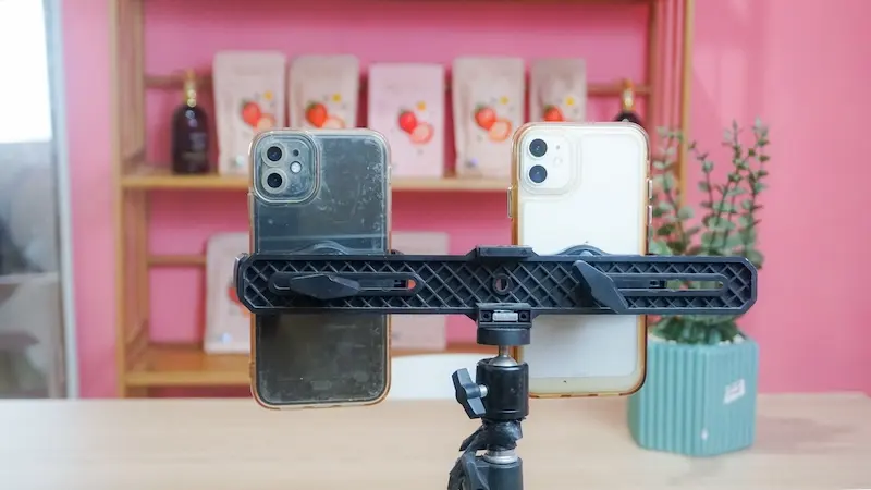 Tampilan depan dua ponsel pada dudukan horizontal di atas tripod, dengan rak produk dan tanaman hias di latar merah muda.