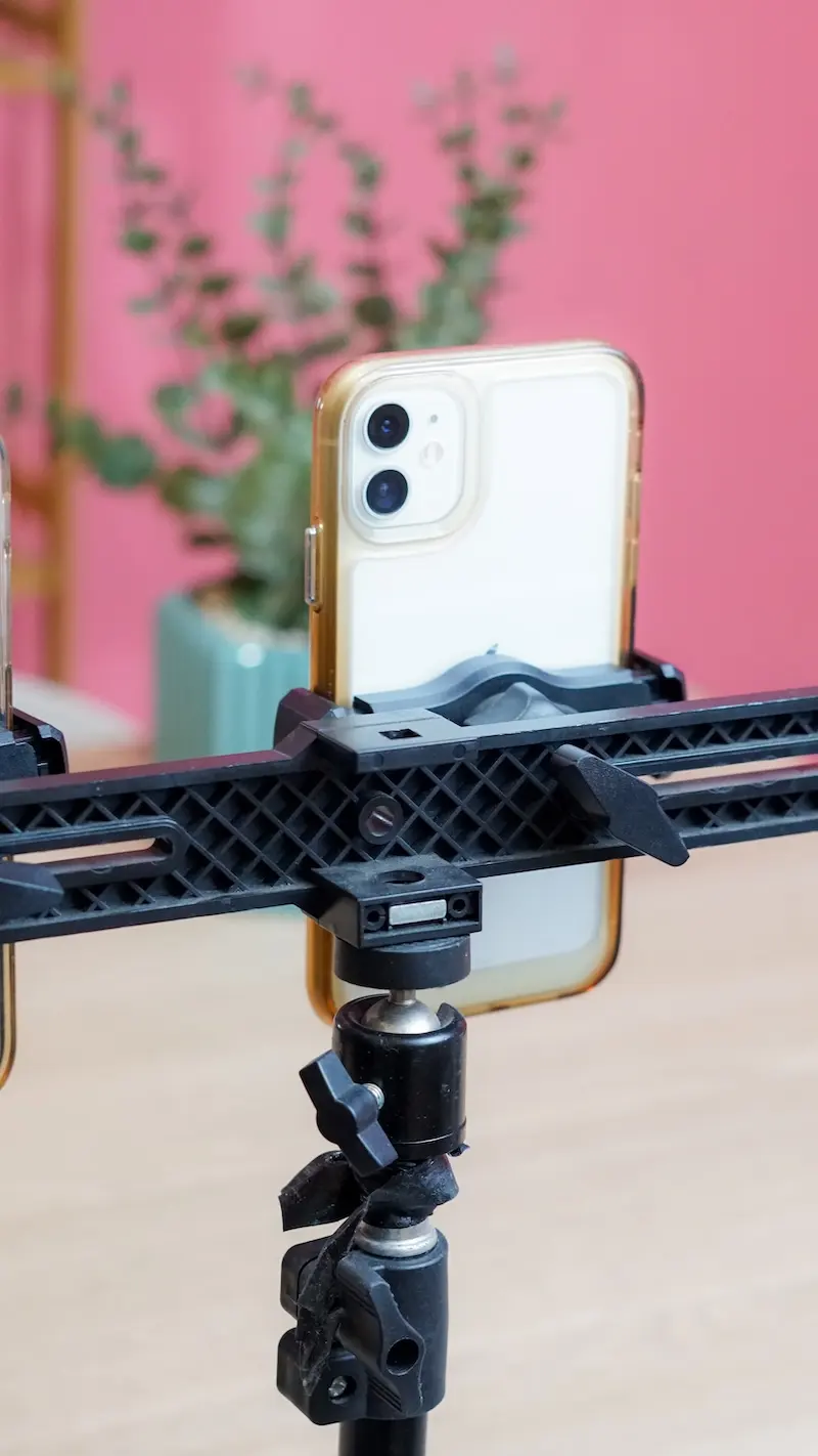 Foto vertikal dekat ponsel putih pada dudukan horizontal hitam dan kepala tripod, berlatar studio merah muda.