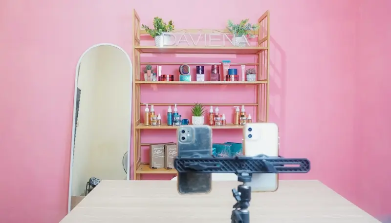Studio merah muda dengan meja, rak produk, dan dua ponsel pada dudukan.