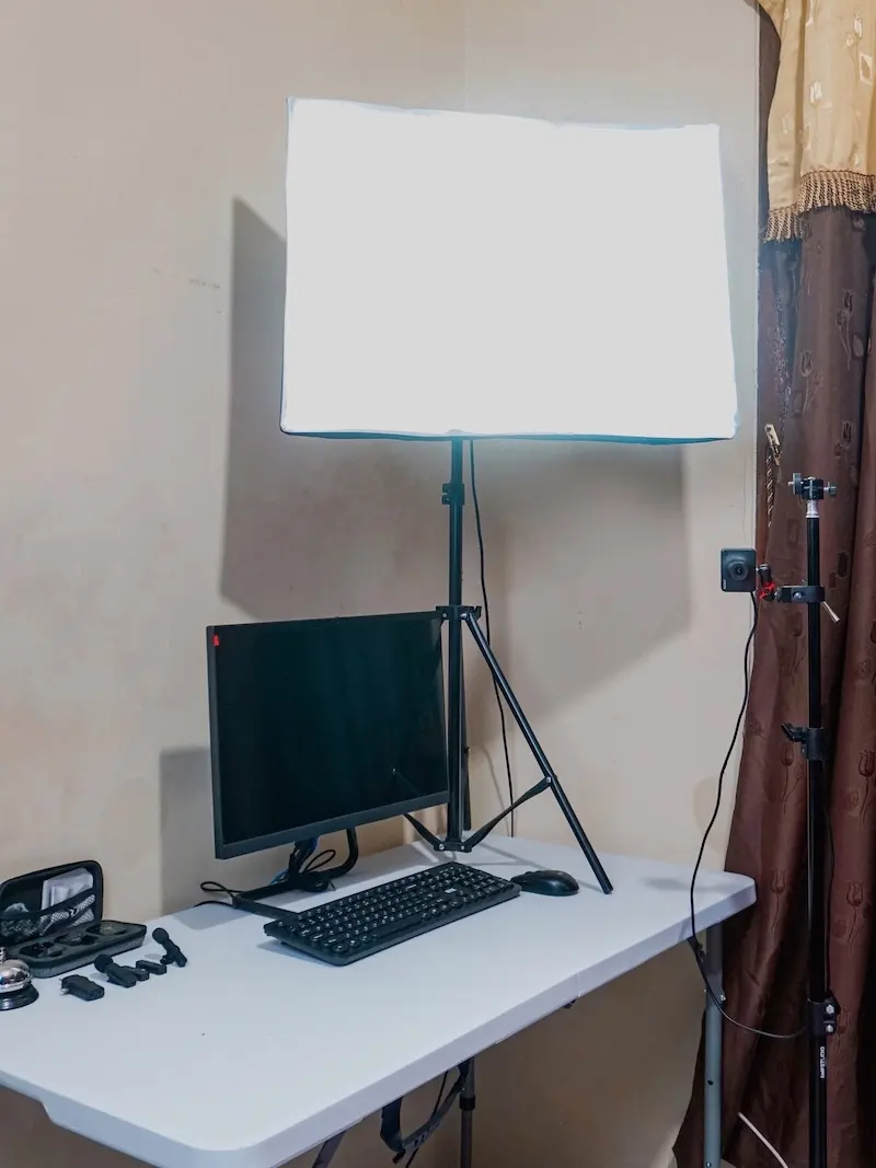 Lampu softbox persegi menyala di atas monitor gelap dan keyboard pada meja putih, di samping tirai cokelat.